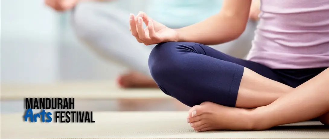 two women sit cross-legged on yoga mats, wearing light-coloured clothing, with their hands resting on their knees